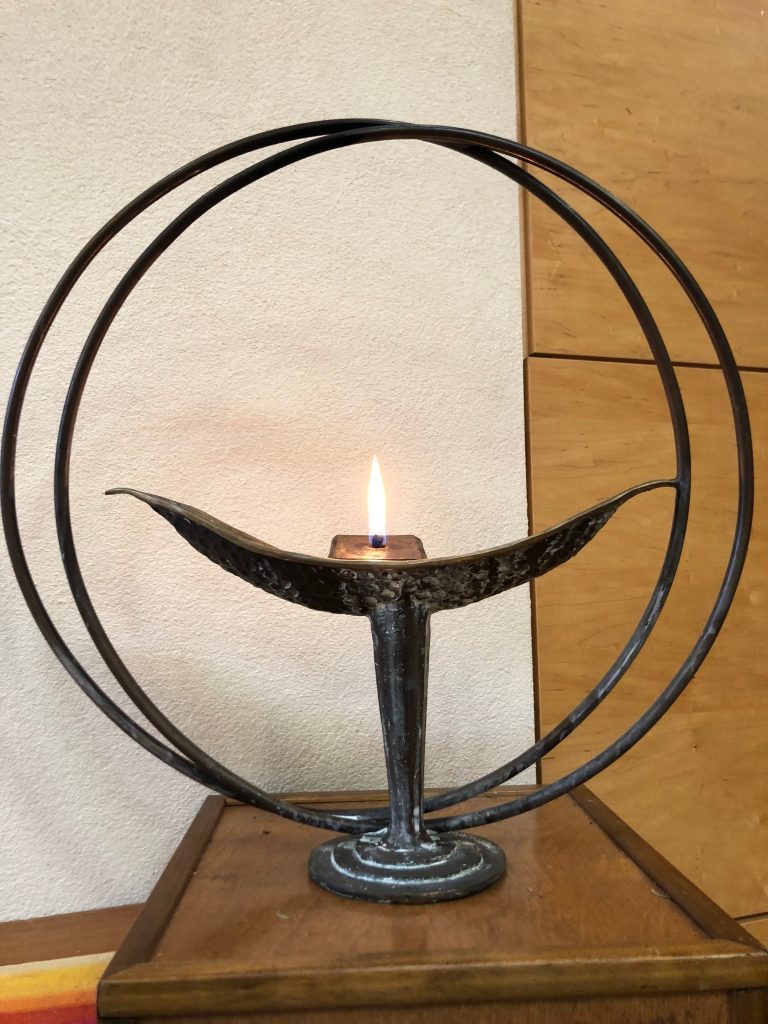 Delicate black metal chalice with lighted white candle within two large metal circles, against beige and wood background.